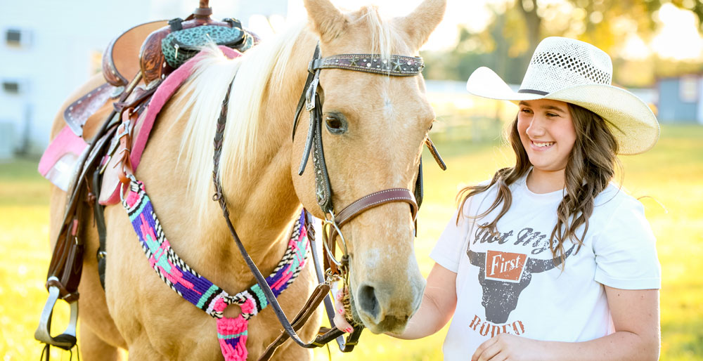 Ella Nipper standing next to her palomino horse, wearing a straw cowboy hat and smiling.